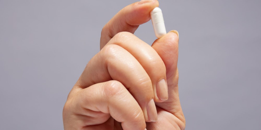 Hand holding a white capsule pill against a neutral background.