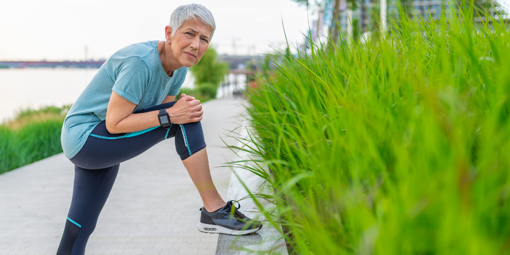 Older woman in workout clothes stretching outdoors by tall grass.